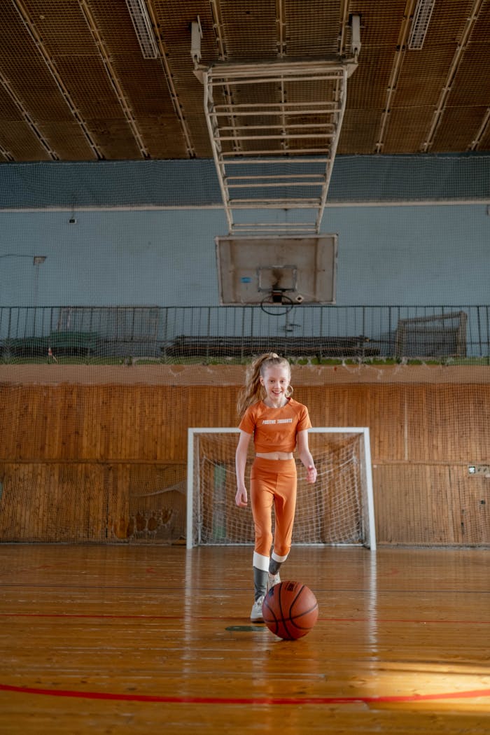 Girl in an orange outfit dribbling a basketball indoors, joyful and active.